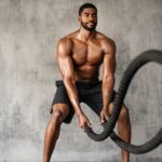 Man with no shirt on working out with battle ropes.