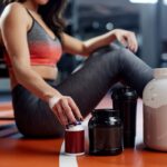 Woman sitting on gym floor with various unlabeled fitness supplements.