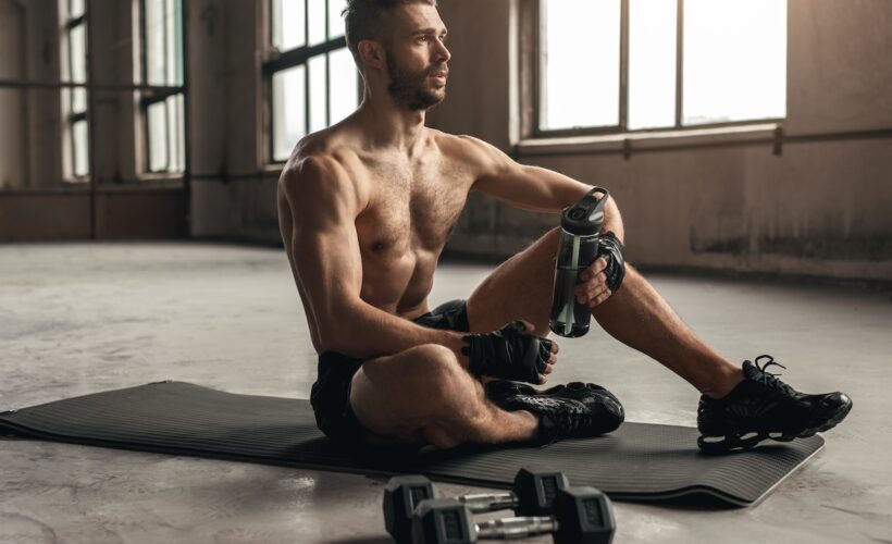 Strong male athlete with bottle of water looking away while resting on mat in gym.