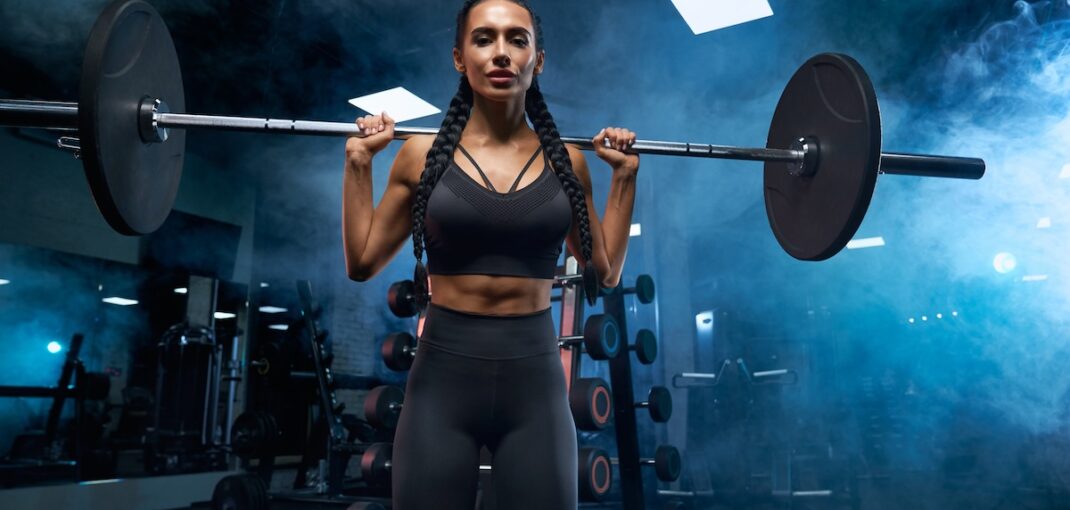 Woman with long braids holding barbell on shoulders in gym.
