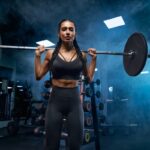 Woman with long braids holding barbell on shoulders in gym.