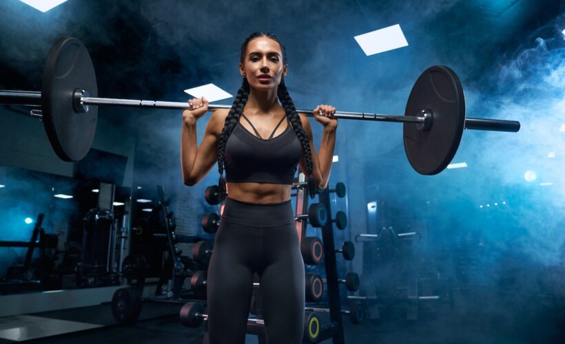 Woman with long braids holding barbell on shoulders in gym.