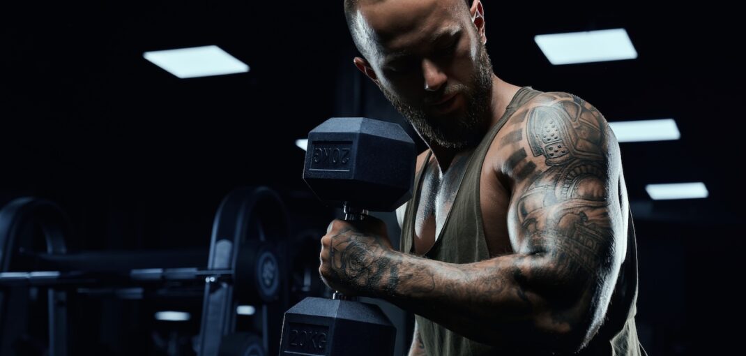 Side photo of tattooed weightlifter performing bicep curls with a dumbbell.